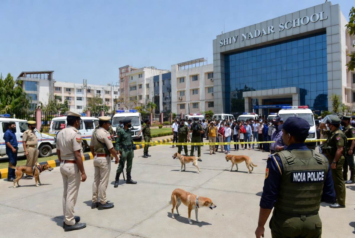 A realistic photojournalistic news image of Noida police and bomb disposal squad (BDS) with sniffer dogs searching a modern school campus in Noida, India on February 19 2026 during CBSE board exams. Students and parents standing outside in safe zone, evacuated, worried faces, police vans and security tape around the school building, clear sunny day, high detail, professional news photography style like Reuters or PTI, wide angle shot showing school name board partially visible like ‘Shiv Nadar School’ or ‘DPS’, red alert atmosphere but no actual explosionlandscape